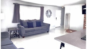 A living room with a grey sofa, a small side table, a large mirror, and a window with curtains.