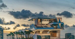 Un hôtel moderne situé au bord de l’eau, doté d’une architecture originale, de plusieurs niveaux et de balcons, et situé à proximité d’une jetée bordée de palmiers.