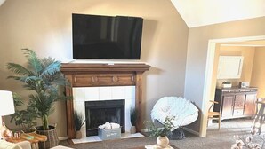 A living room with a fireplace, a television mounted above it, a sofa, a coffee table, and a dining table with a vase of flowers. - Spacious 4 BDRM, Game Room, 85” TV, Fenced Yard (Overland Park)