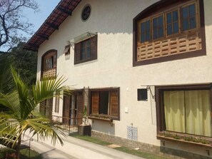 A two-story building with multiple windows, a balcony, and a small garden area with a palm tree. - Pousada Recanto das Marias (Santo Antonio do Pinhal)