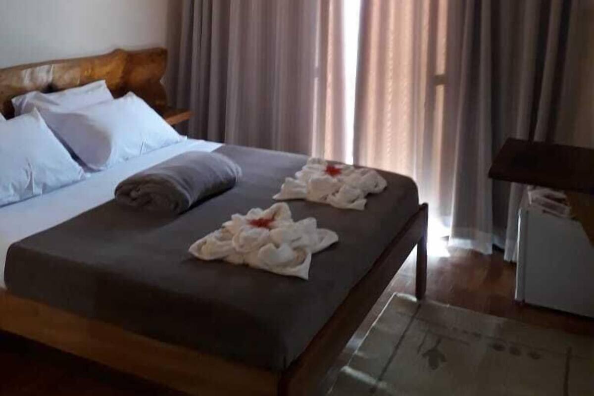 Quarto de hotel com cama, mesa de cabeceira, luminária e janela com cortinas.