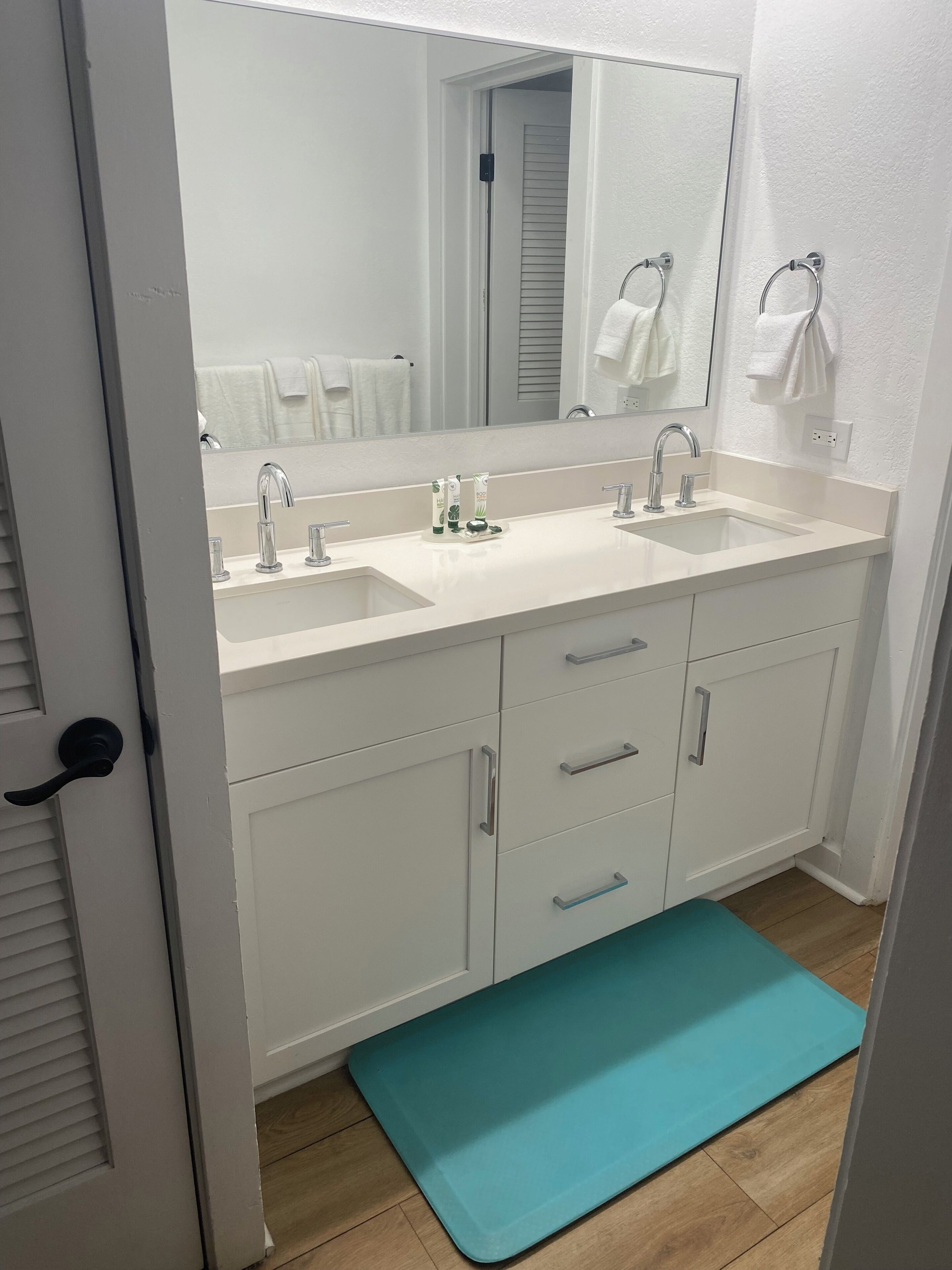 A bathroom with two sinks, a large mirror, and a teal bath mat.