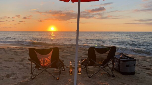 A beach scene with two chairs under a red umbrella, a cooler, and bottles on the sand. - Casa Aerie – Beach Days, Rooftop Nights (San Francisco)