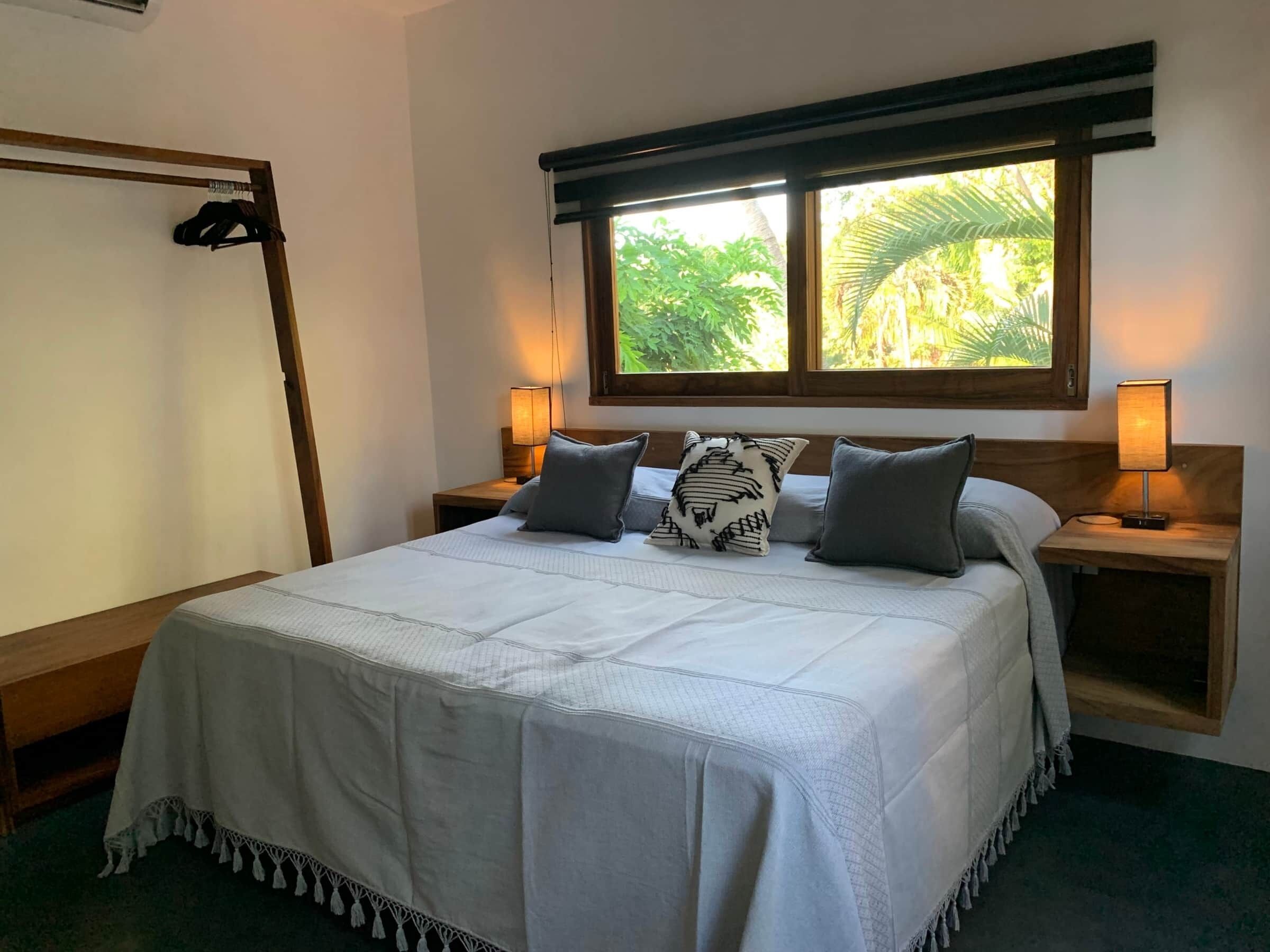 A neatly made bed with a white bedspread and two pillows, flanked by wooden nightstands with lamps, in a room with a large window overlooking greenery.