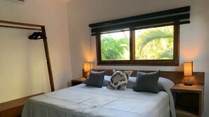 A neatly made bed with a white bedspread and two pillows, flanked by wooden nightstands with lamps, in a room with a large window overlooking greenery. - Casa Aerie – Beach Days, Rooftop Nights (San Francisco)