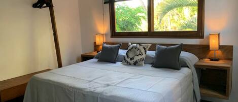 A neatly made bed with a white bedspread and two pillows, flanked by wooden nightstands with lamps, in a room with a large window overlooking greenery.