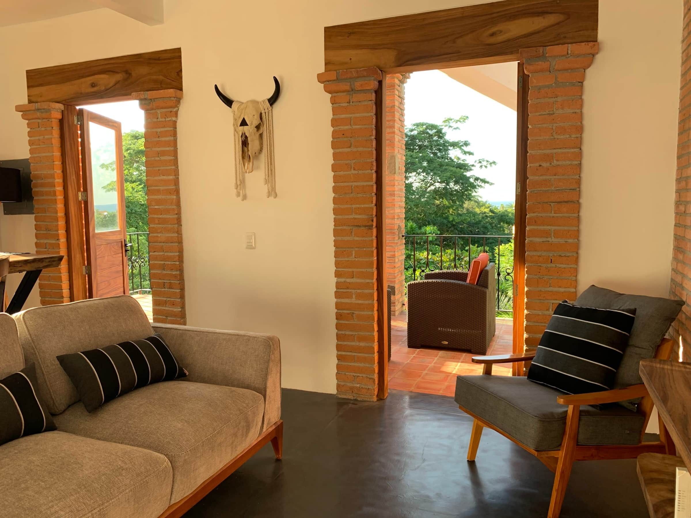 A living room with a sofa, a chair, and a decorative skull on the wall. There are open doors leading to an outdoor area with a balcony.