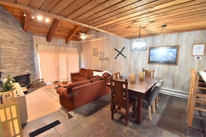 A cozy living room with a stone fireplace, wooden ceiling beams, a sofa, a dining table with chairs, and a mounted TV.
