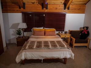 A cozy bedroom with a wooden ceiling, a bed with patterned bedding, a nightstand with a lamp, a chair, and a teddy bear.