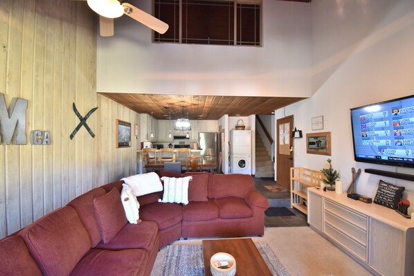 Una sala de estar moderna con un sofá modular, un televisor de pantalla plana y una cocina al fondo.