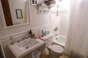 Un baño con lavamanos, inodoro y bañera blancos. Hay un perchero sobre el lavamanos y una cortina de ducha al fondo.