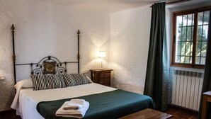 A neatly made bed with a green bedspread, a wooden bench, a bedside table with a lamp, and a window with curtains. - Rural house with patio and garden (Alcalá la Real)