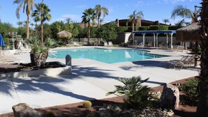 Un área de piscina con sillones de descanso, palmeras y una zona de estar cubierta.