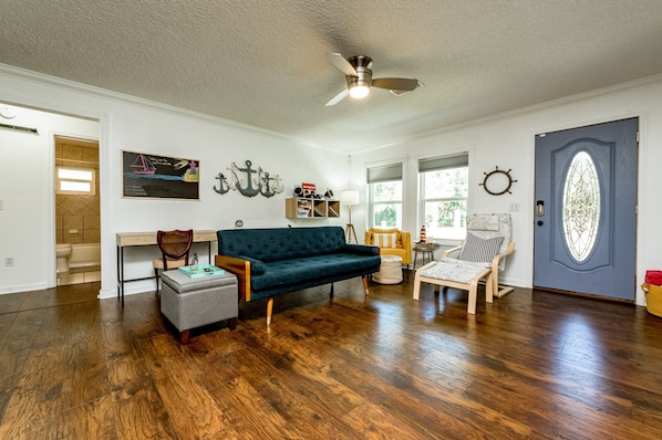 Uma sala de estar ampla com piso de madeira, um sofá azul escuro, uma mesa de centro de madeira e uma pequena mesa lateral. Há um ventilador de teto, uma estante de livros e uma porta com painel de vidro decorativo.