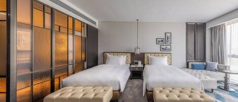 Habitación de hotel con dos camas, un ventanal con vistas, una mesita de noche y un rincón de estar con un sillón y reposapiés.