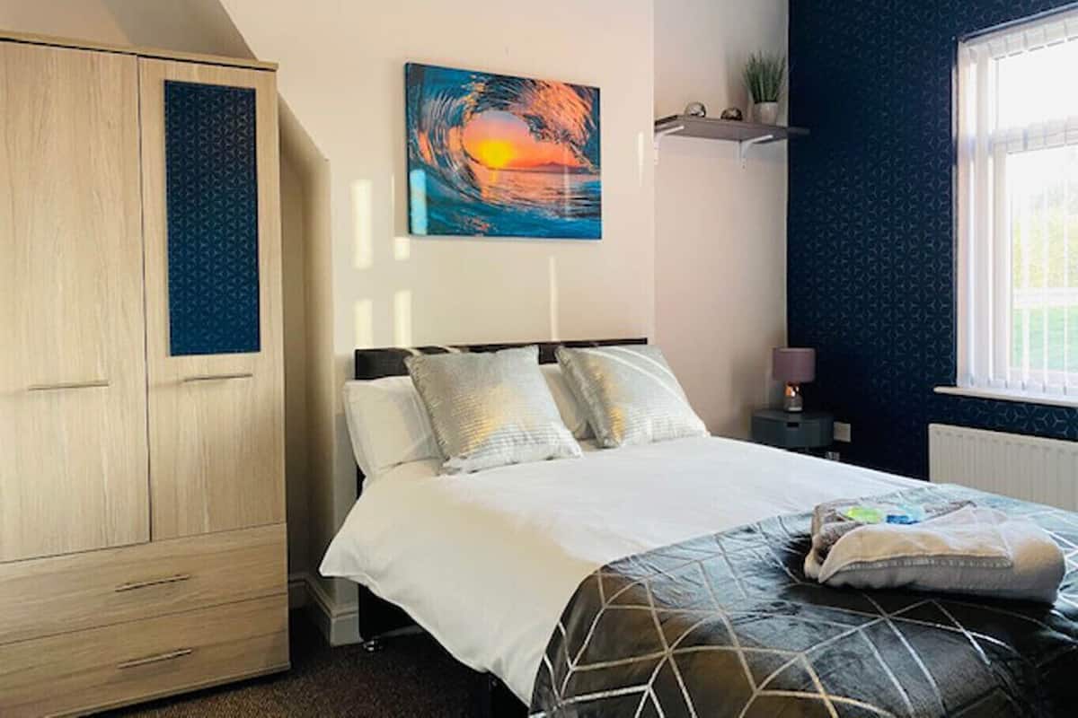 Un dormitorio con cama, armario, un cuadro, ventana y mesita de noche.
