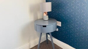 A corner of a room with a blue patterned wall, a grey side table with a lamp, and a power outlet. - Townhouse @ 110 Etruria Vale Road Stoke (Stoke-on-Trent)