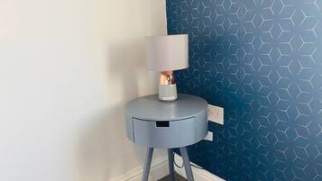 A corner of a room with a blue patterned wall, a grey side table with a lamp, and a power outlet.