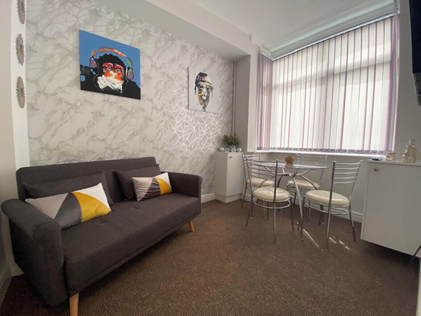 A modern living room with a grey sofa, a dining table with chairs, and a window with blinds.