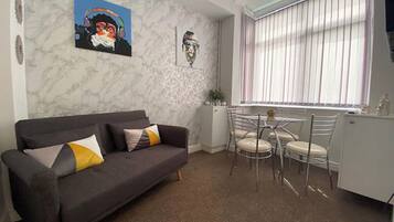 A modern living room with a grey sofa, a dining table with chairs, and a window with blinds.