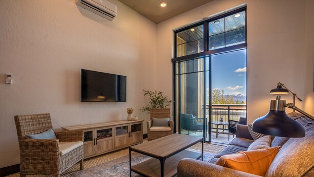 A living room with a large window, a flat-screen TV, a sofa, a coffee table, and a wicker chair. - Upscale Lakeview Condo in Downtown Sandpoint (Sandpoint)