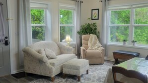 A cozy living room with a floral sofa, a chair with a blanket, a small table, and a view of greenery through large windows. - Mountaintop suite, mountain views, one level, King bed, pet friendly, accessible (Rosman)