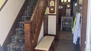 Un escalier recouvert d’une moquette à motifs, un banc en bois, un lustre, une horloge murale et une porte sur laquelle sont suspendus des vêtements.