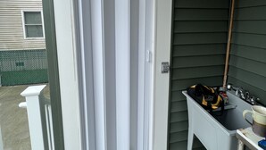 Un espace extérieur abrité, avec une porte blanche, une petite table équipée d’un évier et, au fond, une clôture verte.