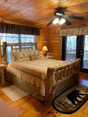 A wooden cabin bedroom with a bed, a ceiling fan, a nightstand, a lamp, and a rug with a bear design.