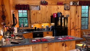 Een eenvoudige keuken in een landelijke hut, met houten kastjes, een centraal eiland met krukken, een koelkast en diverse keukenapparaten.