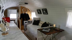 Habitación compacta y bien iluminada con cama, una mesita pequeña y una zona de estar. Una ventana permite la entrada de luz natural, y se observa un electrodoméstico rojo al fondo.