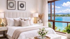 Un dormitorio con cama, mesitas de noche, mesilla de noche, ventilador de techo, ventana con vista al mar y una ventana grande con cortinas.