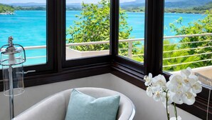 Una habitación con vista al mar turquesa, una silla blanca con cojín azul y un jarrón con flores blancas.