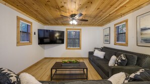 A living room with a wooden ceiling, a ceiling fan, a flat-screen TV, and a dark-colored sofa with patterned cushions.