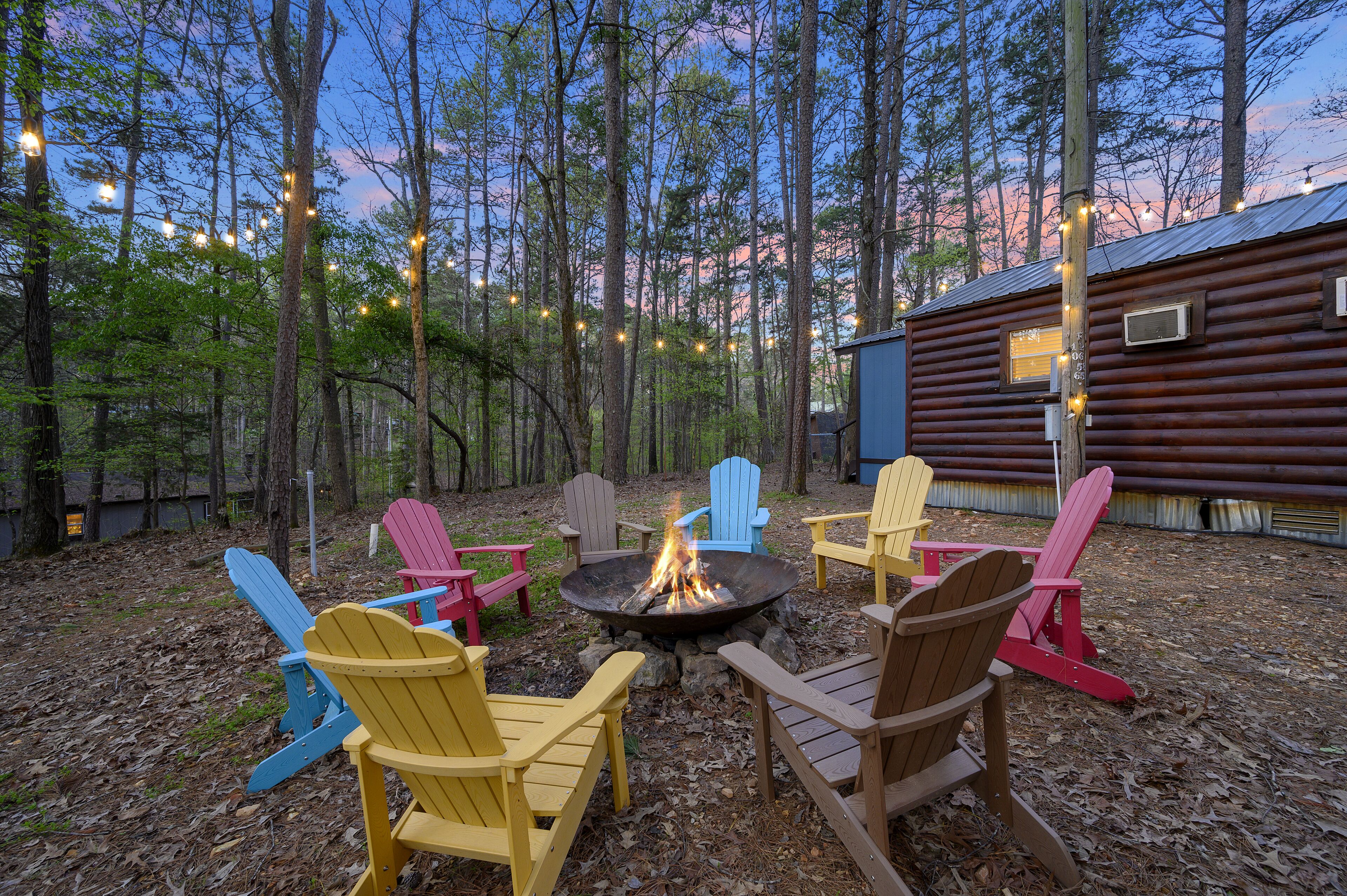 A campfire surrounded by colorful Adirondack chairs in a forest setting with a cabin in the background.