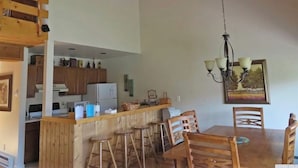 Cuina amb armaris de fusta, zona de menjador amb taula i cadires de fusta, i llum penjant.