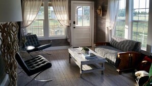 A cozy room with a wooden floor, a white door, a black leather chair, a patterned sofa, and a wooden coffee table. - Molly Cabin  - Private cabin with 2 bedrooms, one queen and one king bed, full bath with walk-n-shower. Full kitchen with fridge/ice maker, coffee bar, front and back porches, private out door fire pit, outdoor shower. (Round Top)