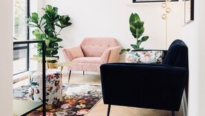 Un salon moderne avec un canapé noir, un fauteuil rose et un fauteuil noir. Une grande fenêtre offre une vue sur la verdure à l’extérieur.