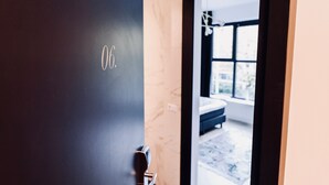 Une chambre d’hôtel avec une porte sombre portant le numéro 06, un lit et une vue sur l’extérieur par une fenêtre.