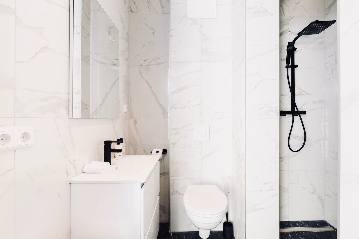 Un baño moderno con un lavamanos de mármol blanco, un inodoro y un cabezal de ducha negro.