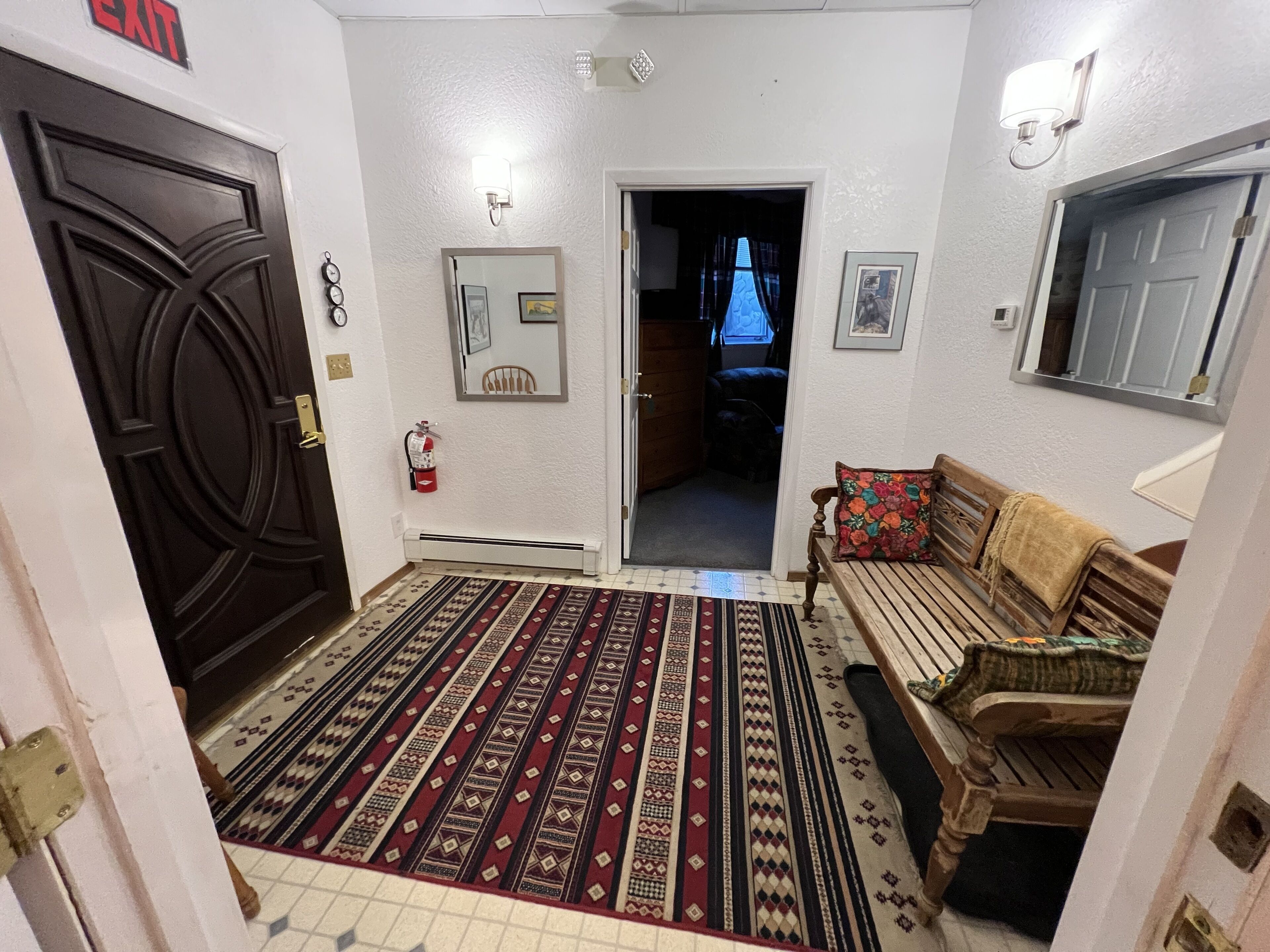 A hallway with a patterned carpet, a wooden bench with a colorful cushion, a door with an 'EXIT' sign, and a wall-mounted light fixture.