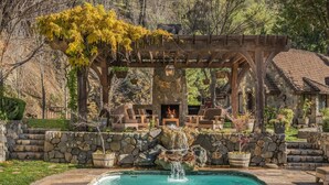 A stone pool with a waterfall, wooden pergola, outdoor seating, and a stone wall.
