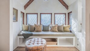 A cozy reading nook with a cushioned bench, a round ottoman, and a wooden ceiling.