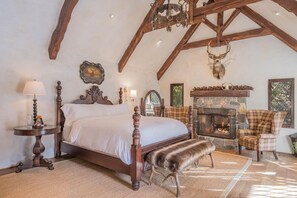 Une chambre avec un grand lit, une cheminée en pierre, une tête de cerf décorative et un fauteuil à motifs à carreaux.