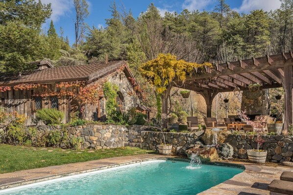 Un chalet rustique avec un mur en pierre, une piscine et une pergola en bois avec des sièges d’extérieur.