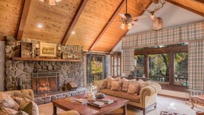 A cozy cabin interior with a stone fireplace, wooden ceiling beams, a plush sofa, and a large window offering a view of the outdoors.
