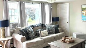 Una sala de estar con sofá, mesa de centro y sillón. Hay un ventilador de techo, una ventana con cortinas y una puerta.