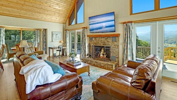 A spacious living room with leather sofas, a fireplace, and large windows offering a view of the outdoors. - Luxury Mountaintop Views with Hot Tub - 1 min to town (Hiawassee)