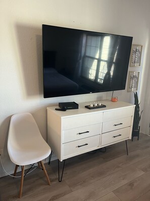 A modern living room with a flat-screen TV mounted on the wall, a white cabinet with drawers, a white chair, and a potted plant.
