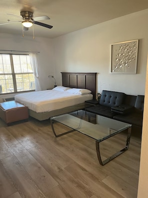 A hotel room with a bed, a glass coffee table, a sofa, a ceiling fan, and a window with curtains.